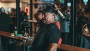 A man is holding an axe over his shoulder with a playful expression, likely at an axe-throwing venue or similar social setting.