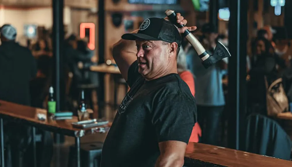 A man is holding an axe over his shoulder with a playful expression likely at an axe-throwing venue or similar social setting