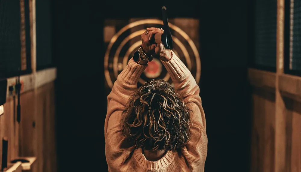 A person is seen from behind preparing to throw an axe at a target in an indoor axe-throwing range