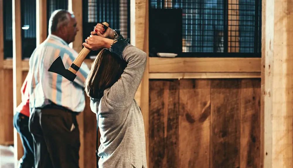 A person is seen from behind holding an axe above their head seemingly about to throw it at a target in an indoor axe-throwing range with another person watching in the background