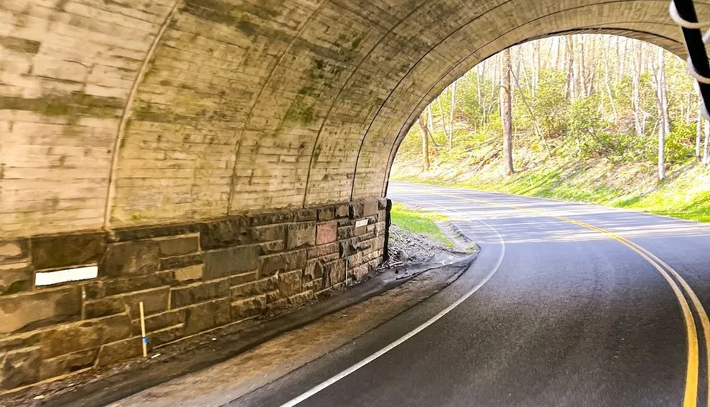 The image shows a road curving to the left as it emerges from a stone tunnel with sunlight filtering in from the forested area outside