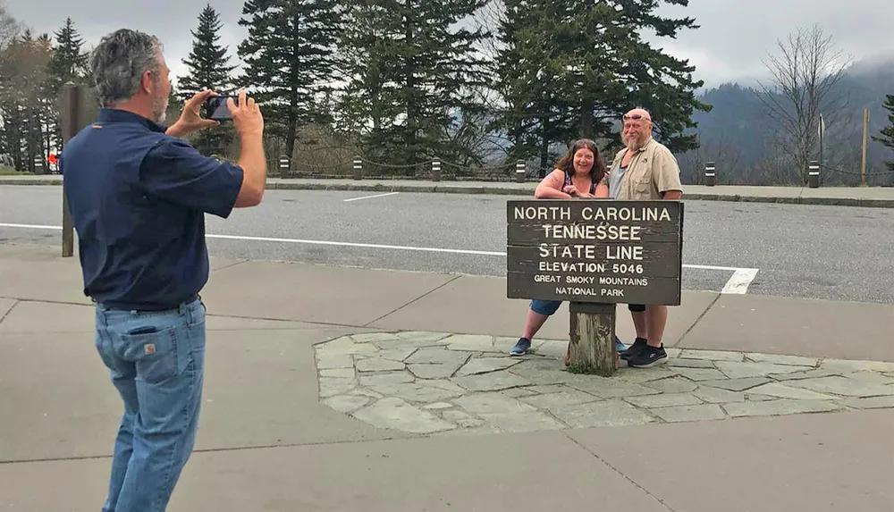 A person is taking a photo of two people standing by a sign marking the state line between North Carolina and Tennessee in the Great Smoky Mountains National Park