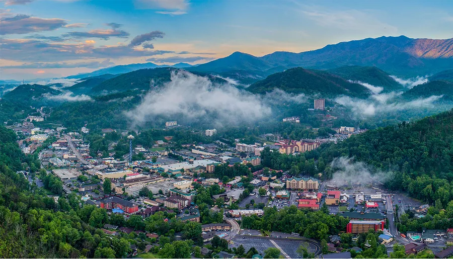 A scenic aerial view captures a town nestled among lush green hills and misty mountains under a partly cloudy blue sky.