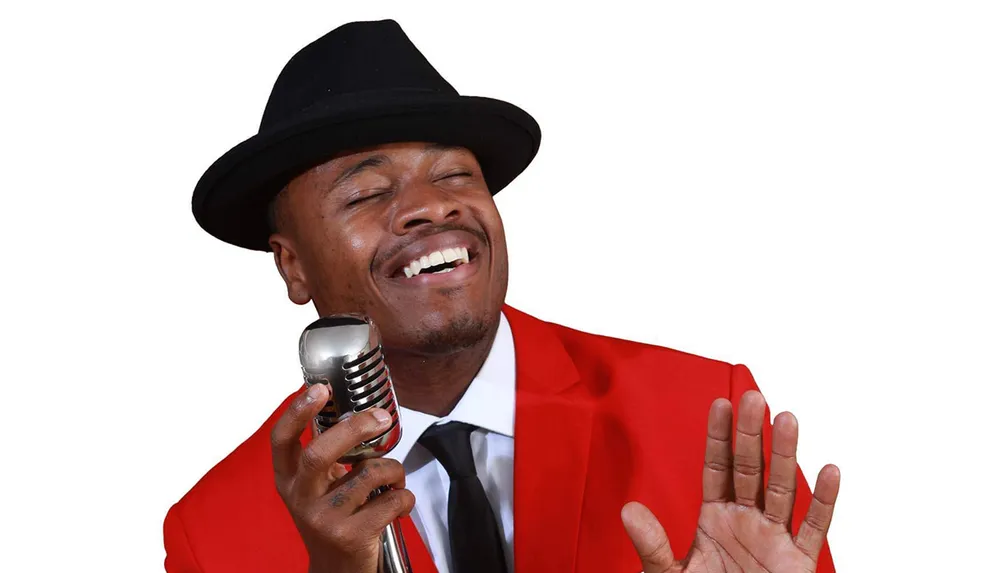A person in a red suit and black fedora is passionately singing into a vintage microphone with a joyful expression
