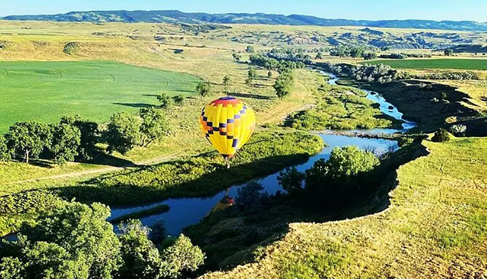 A colorful hot air balloon floats above a winding river in a lush undulating landscape