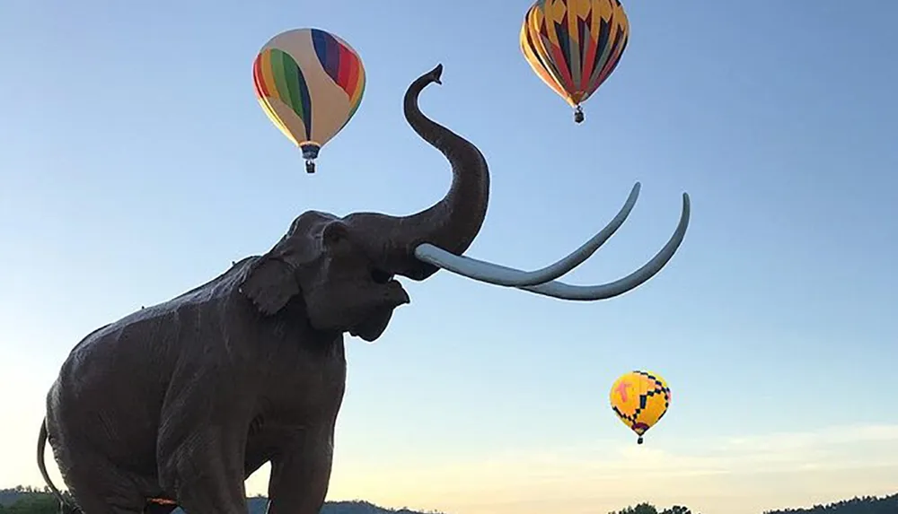 A statue of a mammoth with long tusks is poised against a sky with hot air balloons floating in the background