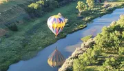A colorful hot air balloon floats above a serpentine river, casting a reflection on the water as it flies over a lush landscape.