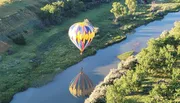 A colorful hot air balloon floats above a serpentine river, casting a reflection on the water as it flies over a lush landscape.