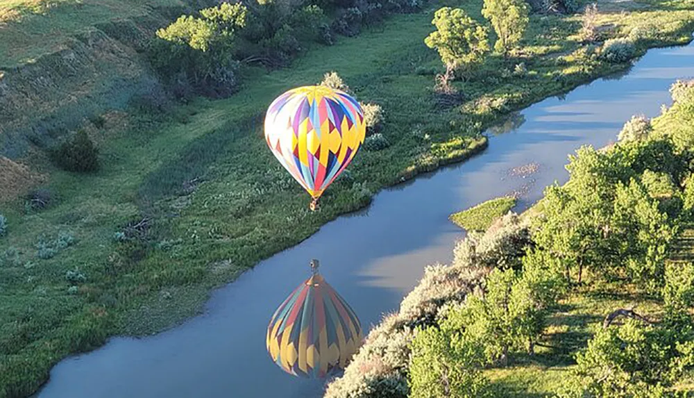 A colorful hot air balloon floats above a serpentine river casting a reflection on the water as it flies over a lush landscape