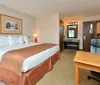 The image shows a neatly arranged hotel room with a large bed a work desk and chair an open bathroom vanity area and warm inviting lighting