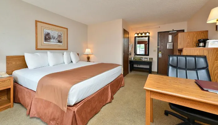 The image shows a neatly arranged hotel room with a large bed a work desk and chair an open bathroom vanity area and warm inviting lighting