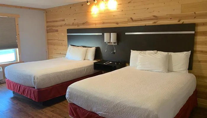 The image shows a neatly arranged room with two double beds against a wall featuring a blend of wood paneling and a dark painted section accentuated by modern light fixtures above each bed