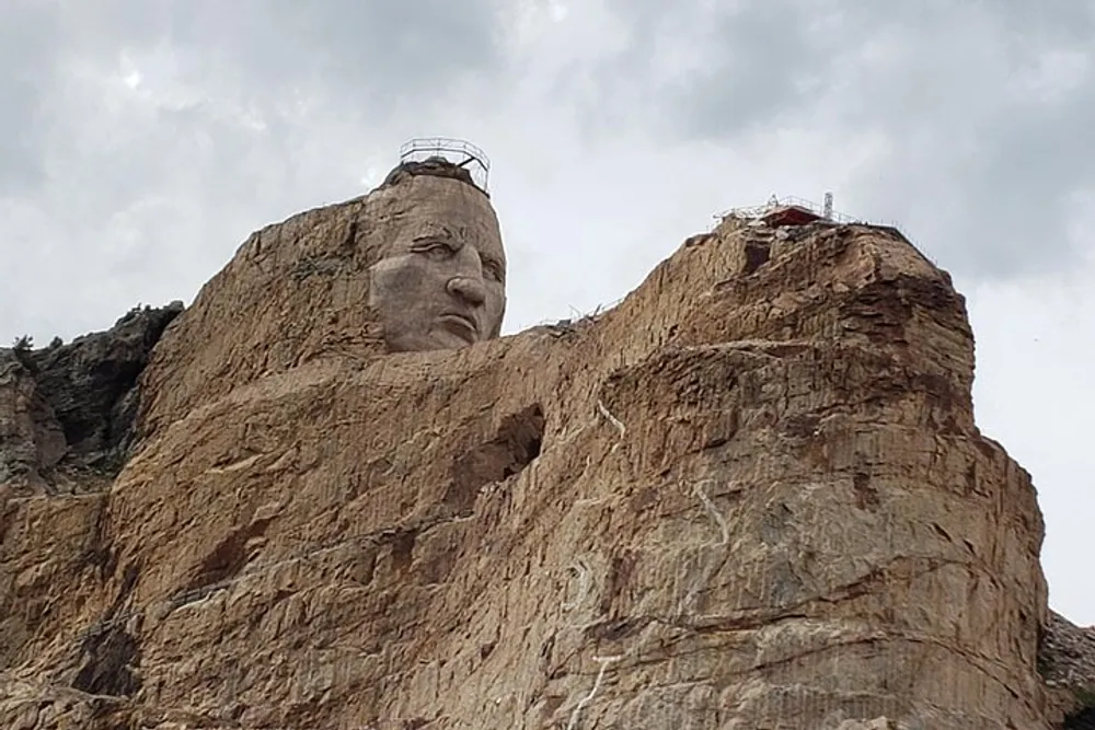 The image shows a massive sculpture of a head carved into the side of a rocky cliff under a cloudy sky