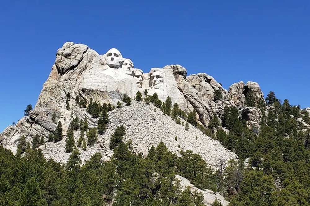 The image shows the iconic Mount Rushmore National Memorial with the carved faces of four American presidents set against a clear blue sky