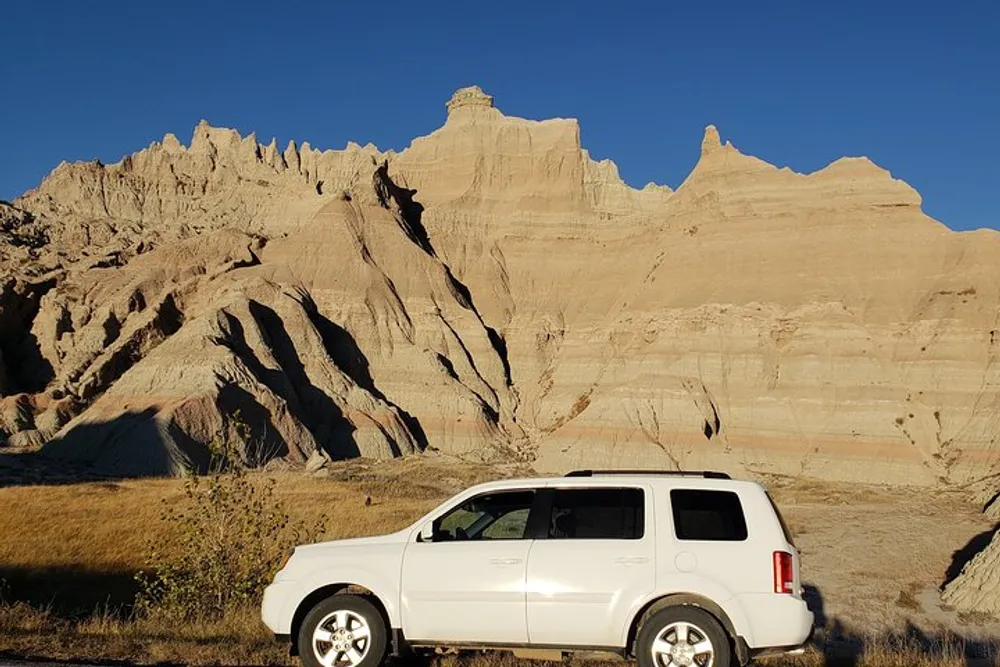 A white SUV is parked in front of a rugged eroded rock formation under a clear blue sky