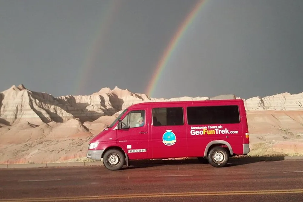 A red tour van is parked on the side of a road with a vivid rainbow appearing in the background over a rocky landscape