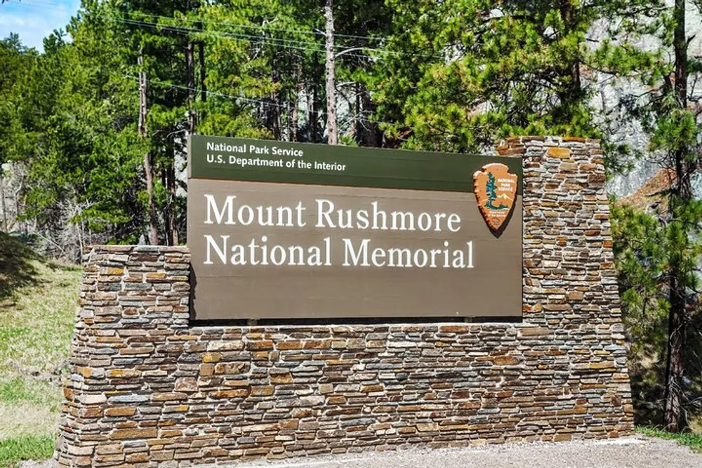 The image shows a sign for the Mount Rushmore National Memorial denoting the entrance to the famous landmark managed by the National Park Service US Department of the Interior