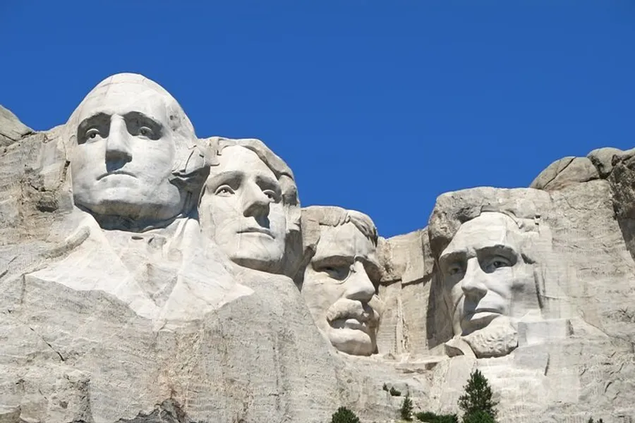 The image shows the iconic Mount Rushmore National Memorial with the carved faces of four American presidents set against a clear blue sky.