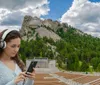 A young woman wearing headphones is focused on her phone in the foreground with the iconic Mount Rushmore National Memorial visible in the background