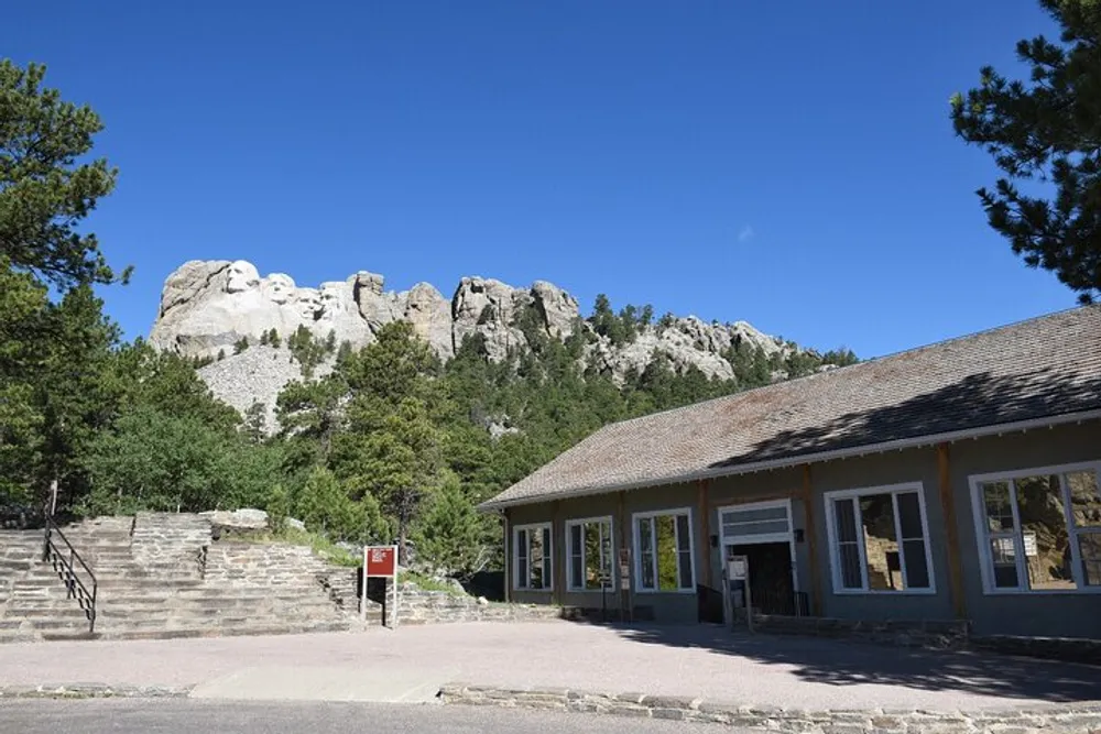 The image shows a building possibly a visitor center with the carved faces of Mount Rushmore visible in the distant background under a clear blue sky