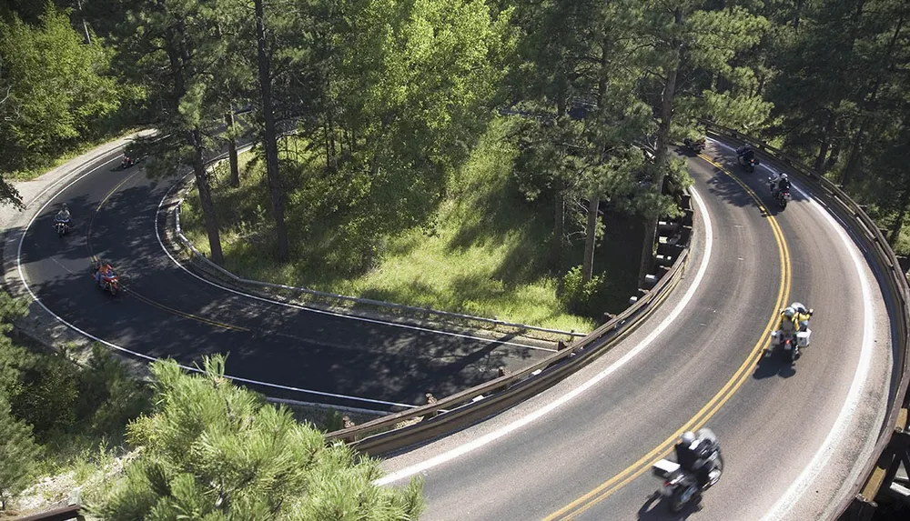 The image shows a winding road through a forested area with multiple motorcycles navigating the curve