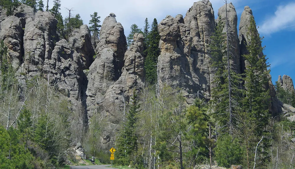 The image depicts a rugged landscape featuring towering rock formations set against a blue sky with coniferous trees and a winding road sign at the base