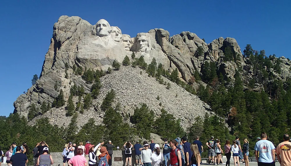 This image shows a crowd of visitors gazing at the iconic Mount Rushmore National Memorial under a clear blue sky