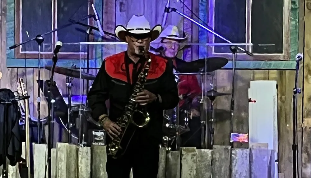 A musician in a cowboy hat is playing the saxophone on a stage with another band member in the background