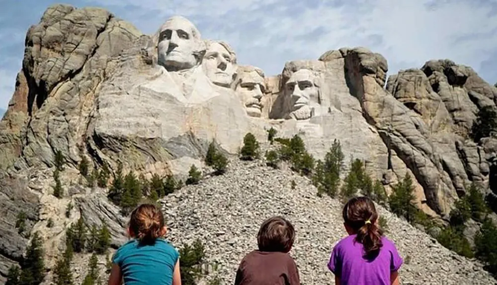 Three individuals are observed from behind gazing upon the iconic Mount Rushmore National Memorial which features the monumental carvings of the faces of four US presidents