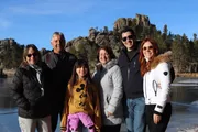 A group of six people, spanning multiple generations, is smiling for a photo in front of a scenic backdrop with rocky formations and a frozen lake.