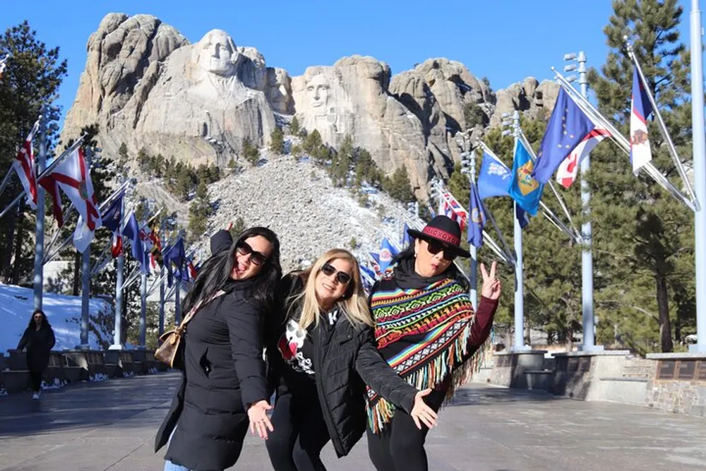Three people are posing for a photo in front of Mount Rushmore under a blue sky with flags on flagpoles in the background