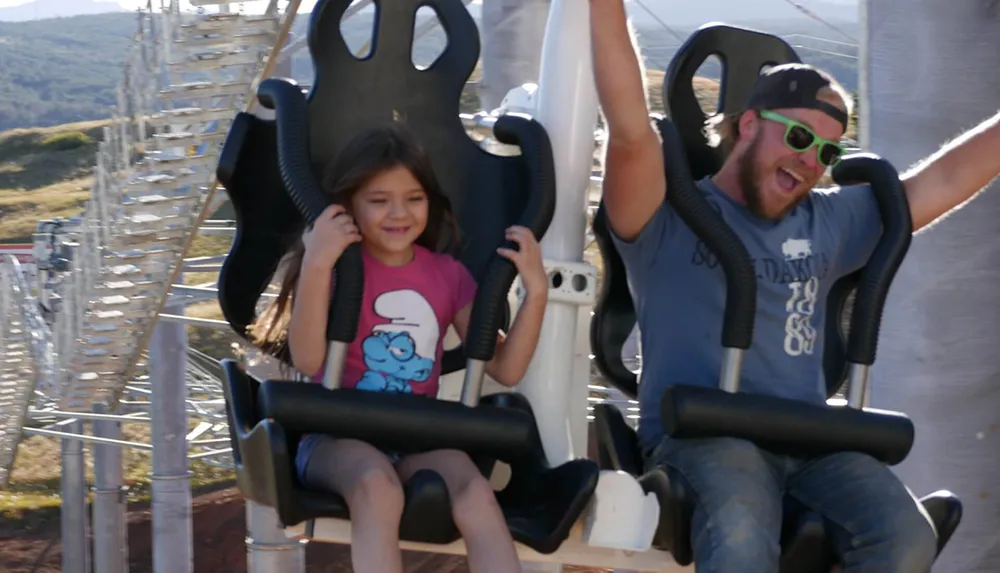A child and an adult are experiencing different reactions while riding a thrilling amusement park attraction with the child appearing apprehensive and the adult showing excitement