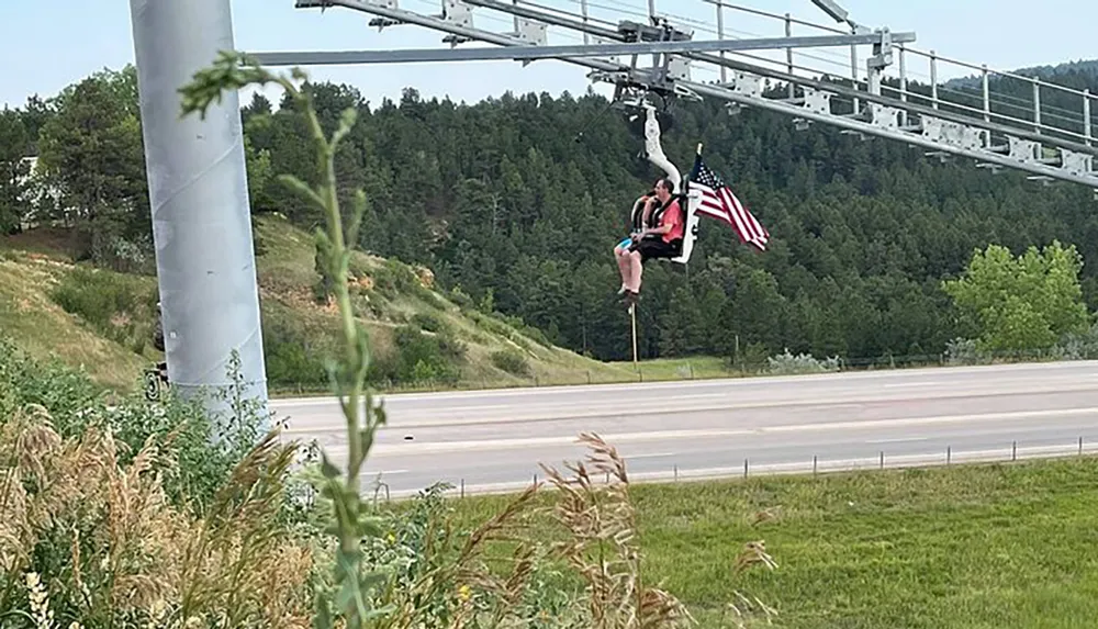 A person is sitting on a chairlift holding an American flag with a scenic backdrop of greenery and a road