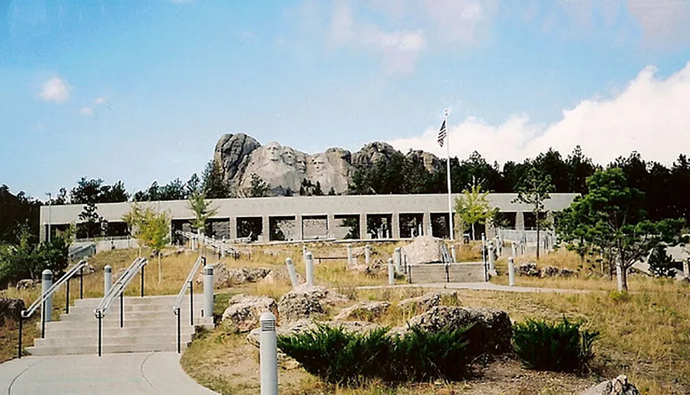 The image shows a clear view of the iconic Mount Rushmore National Memorial in South Dakota with sculptures of four US presidents etched into the granite face of the mountain as seen across the visitor center and an American flag fluttering on the left