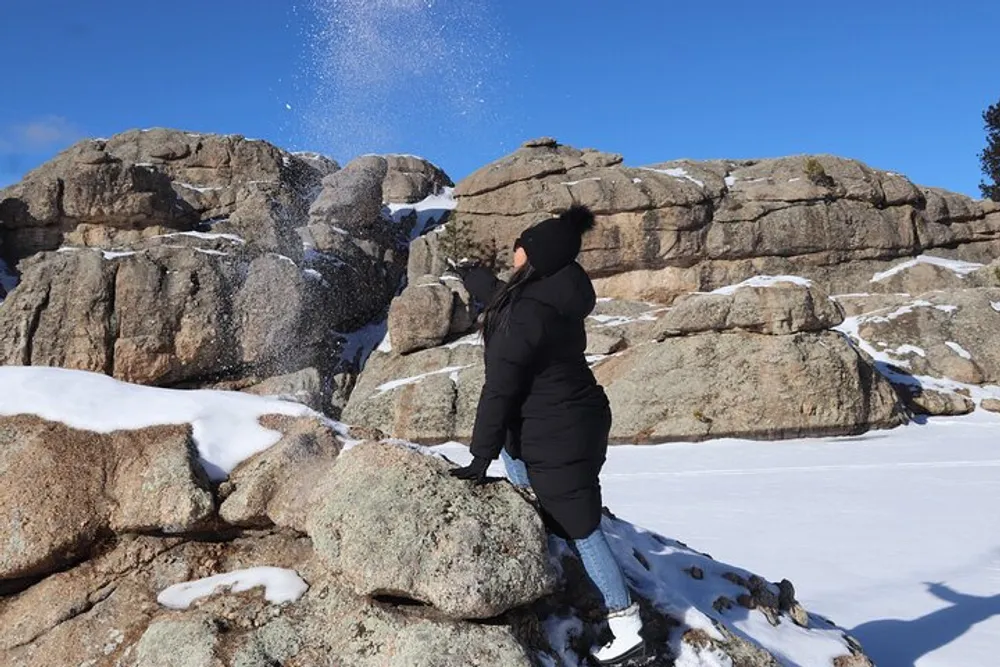 A person in warm winter clothing is throwing snow into the air against the backdrop of a rugged snow-covered rocky landscape under a clear blue sky