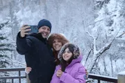 A family of three is taking a selfie together in a snowy environment, each dressed warmly and smiling at the camera.