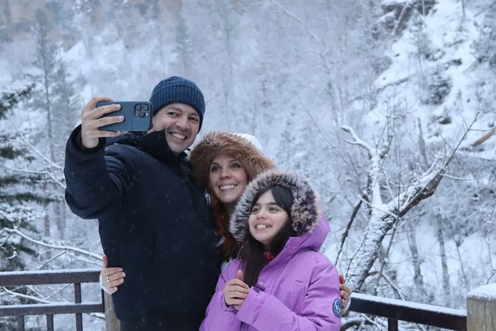 A family of three is taking a selfie together in a snowy environment each dressed warmly and smiling at the camera
