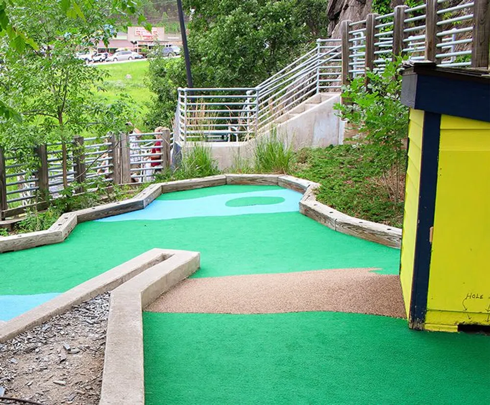 This image shows a colorful outdoor mini-golf course with green artificial turf surrounded by nature and a walkway with metal railings