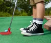 A person wearing black high-top sneakers is playing miniature golf on a green course with a red putter preparing to hit a blue ball