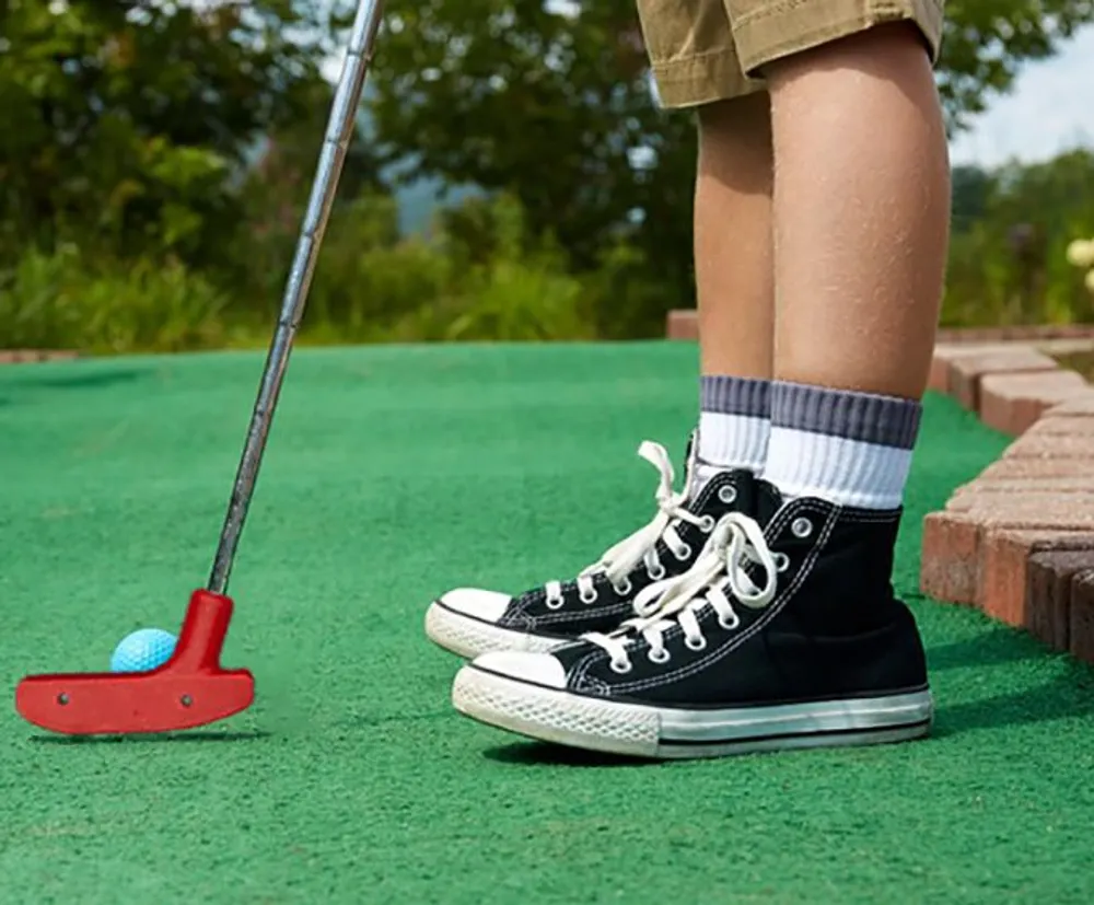 A person wearing black high-top sneakers is playing miniature golf on a green course with a red putter preparing to hit a blue ball