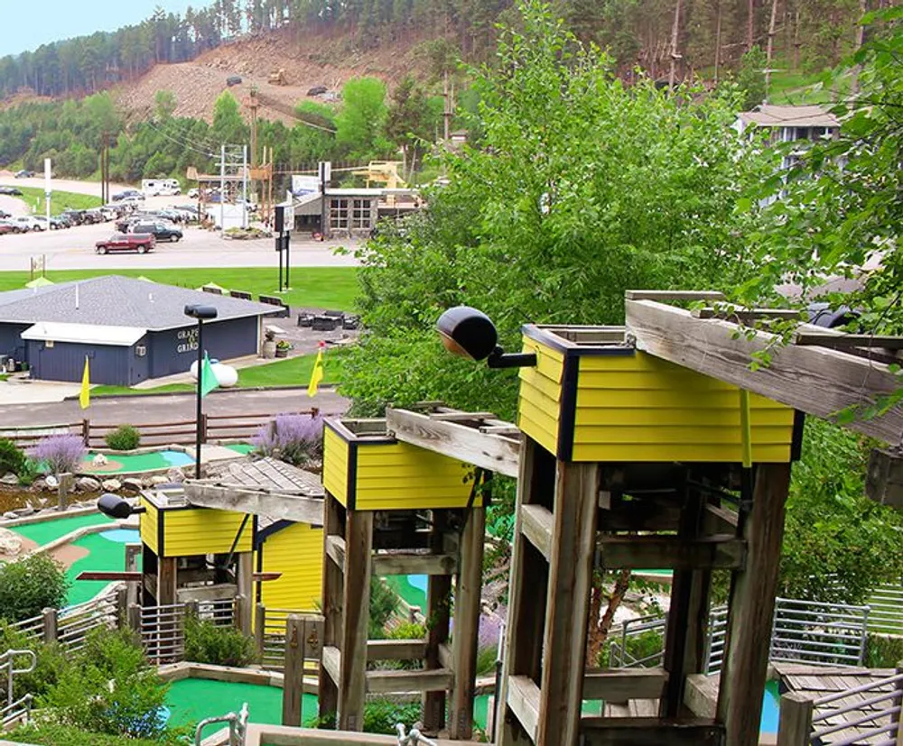 The image shows a colorful outdoor recreational area with a mini-golf course in the foreground and a structure resembling treehouses connected by bridges amidst verdant greenery