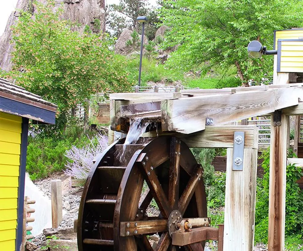 The image shows a wooden water wheel with water flowing onto it surrounded by lush greenery and brightly colored small structures