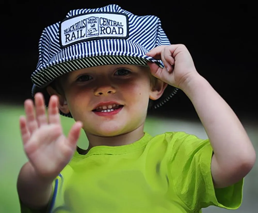 A young child is tipping a striped railroad engineer hat while waving at the camera with a bright smile