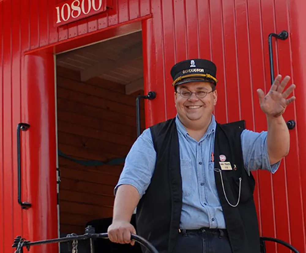 A smiling person in a conductors cap and vest waves from the platform of a bright red caboose