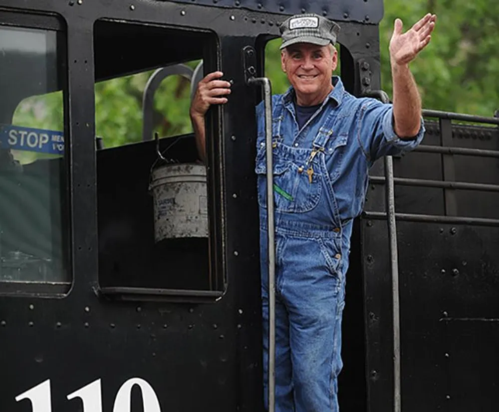 A smiling man in overalls and a cap is waving from the steps of a vintage locomotive