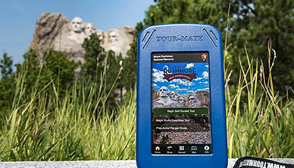 An audio tour device displays options for a Mount Rushmore tour in the foreground with the actual Mount Rushmore monument blurred in the background