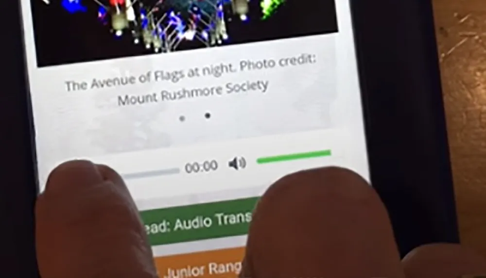 The image shows a close-up view of a persons finger scrolling through a gallery on a digital device that displays a night photo of the Avenue of Flags with a caption crediting the Mount Rushmore Society
