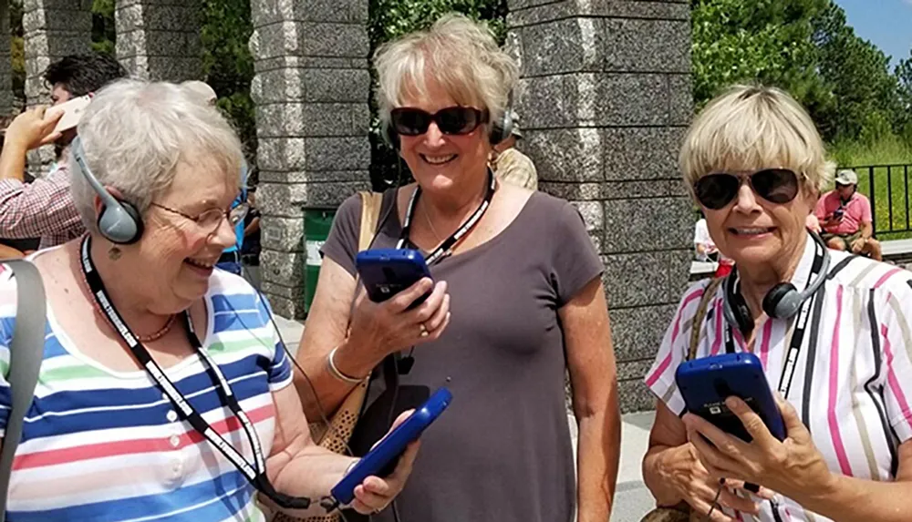 Three smiling women are using audio guide devices likely on an outdoor tour