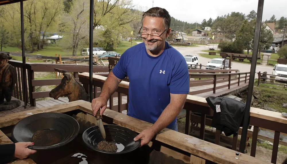 A smiling man is shown panning for gold on an outdoor wooden deck with rustic decor and a view of a rural town in the background