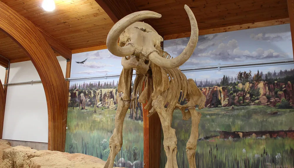 The image shows the skeletal remains of a mammoth on display inside a room with a painted mural depicting a prehistoric landscape